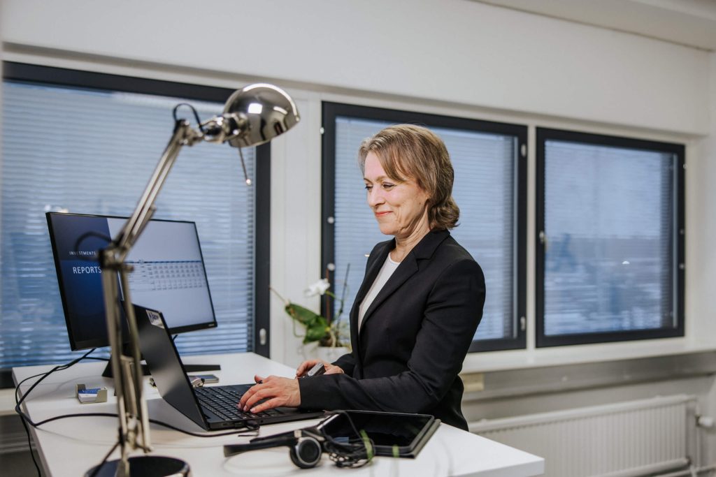 Tiina Hinnen seisoo toimistossa tietokoneen ja tabletin ääressä työskennellen.