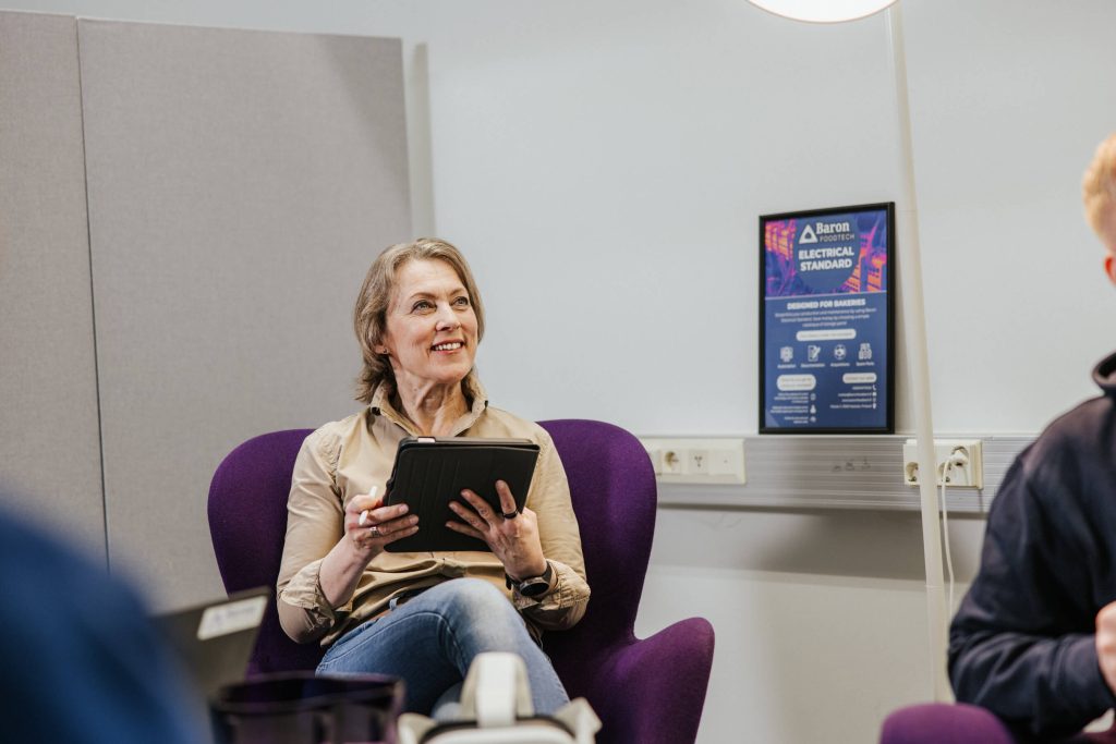 Tiina Hinnen istuu toimistossa violettituolissa ja käyttää tablettia keskustelussa kollegoiden kanssa.