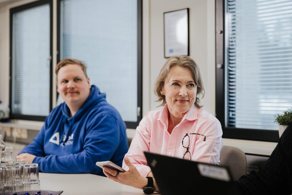 Tiina Hinnen istuu palaverissa vaaleanpunaisessa paidassa ja hymyilee kollegalle.