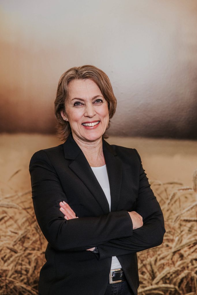 Tiina Hinnen seisoo toimistossa mustassa bleiserissä käsivarret ristissä.