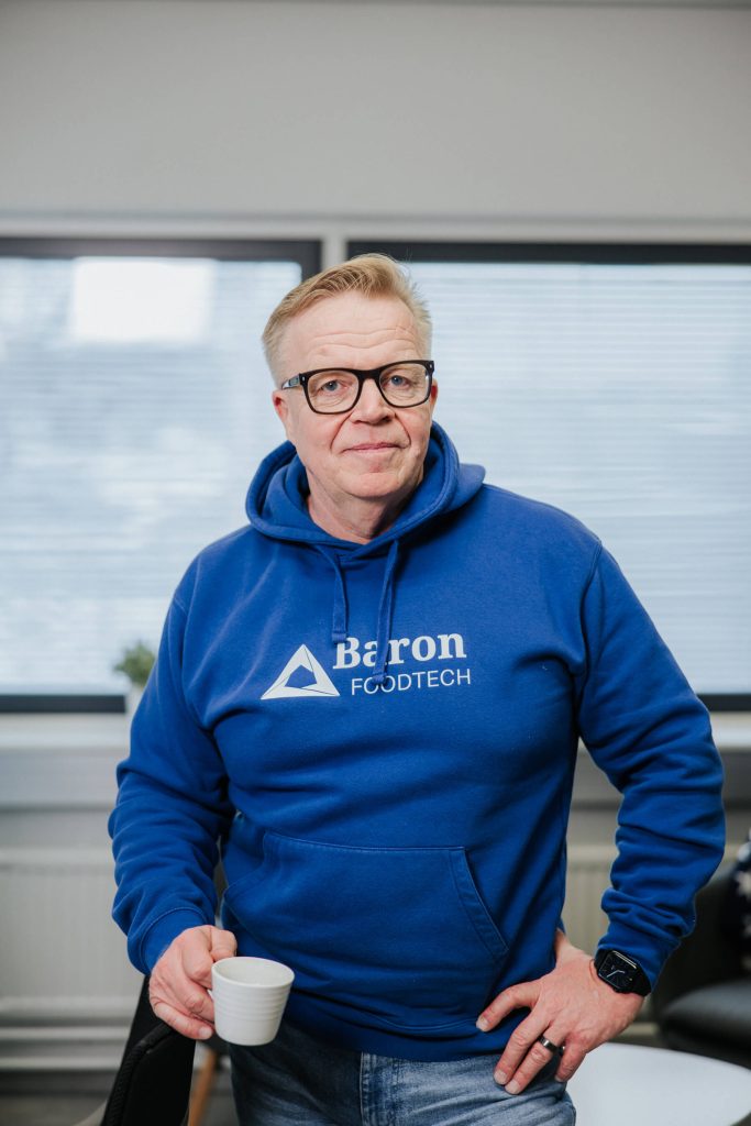 Mikko Lehtinen in a Baron Foodtech hoodie holding a coffee cup and looking toward the camera in an office environment.
